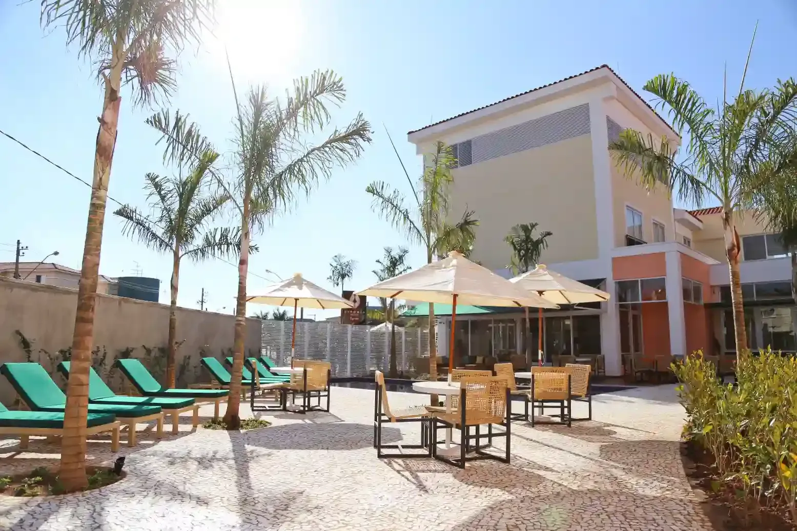 Pátio externo com espreguiçadeiras, mesas e guarda-sóis perto de um prédio e palmeiras sob um céu ensolarado.