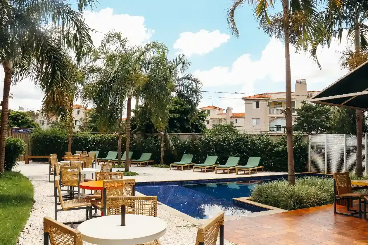 Piscina e área de descanso com mesas, cadeiras e palmeiras sob um céu azul.