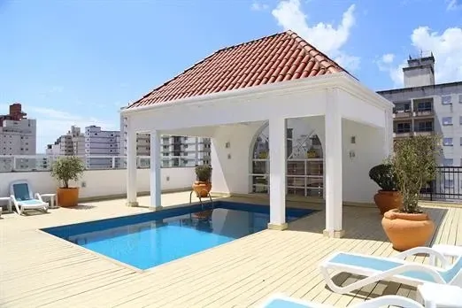 Piscina na cobertura com gazebo branco, água azul e espreguiçadeiras. Dia ensolarado, com a cidade ao fundo.
