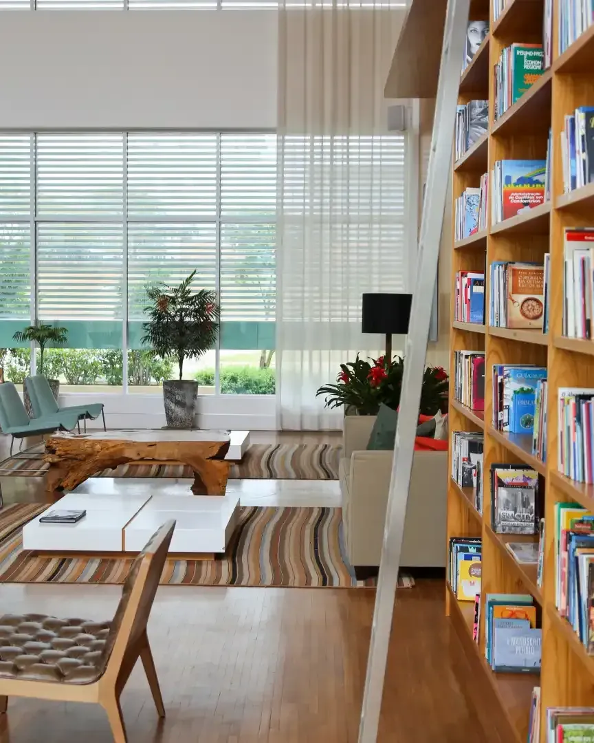 Interior de biblioteca com estante de livros, janelas amplas e assentos.
