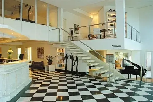 Piso quadriculado em preto e branco em prédio moderno, escada que leva ao segundo andar, piano à direita.