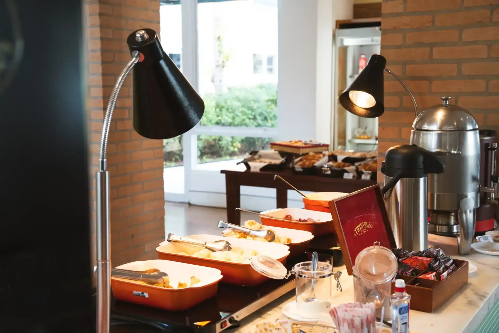 Buffet com exposição de alimentos iluminado por lâmpadas pretas, disposto em frente a uma janela e uma parede de tijolos.