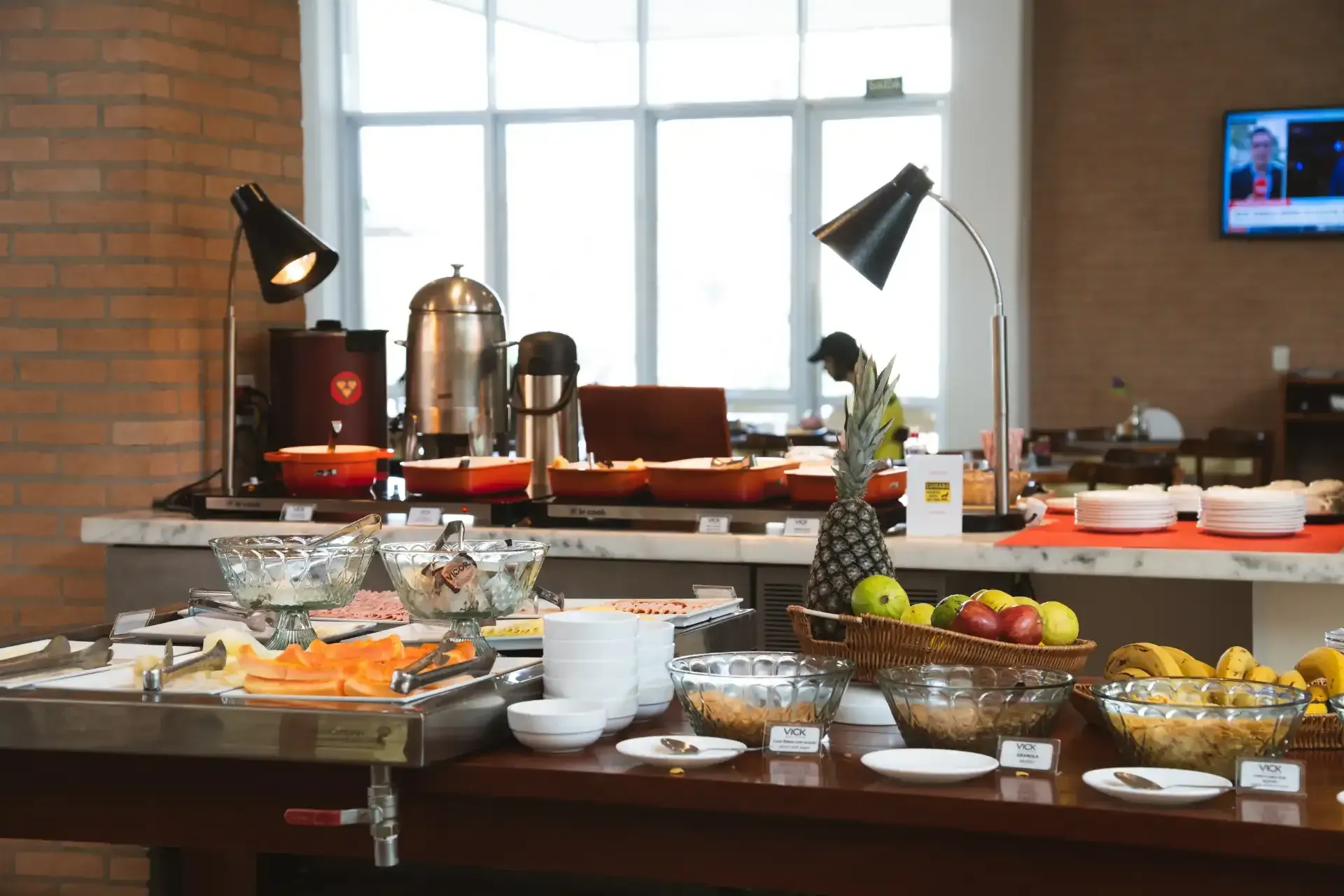 Buffet com exposição de alimentos, incluindo frutas, cereais e pratos quentes, disposto junto a uma janela e uma parede de tijolos.