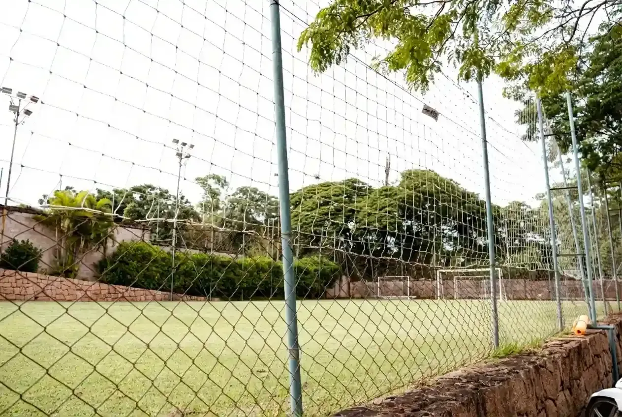 Um campo de futebol gramado cercado por uma cerca de arame, com árvores e um céu nublado ao fundo.