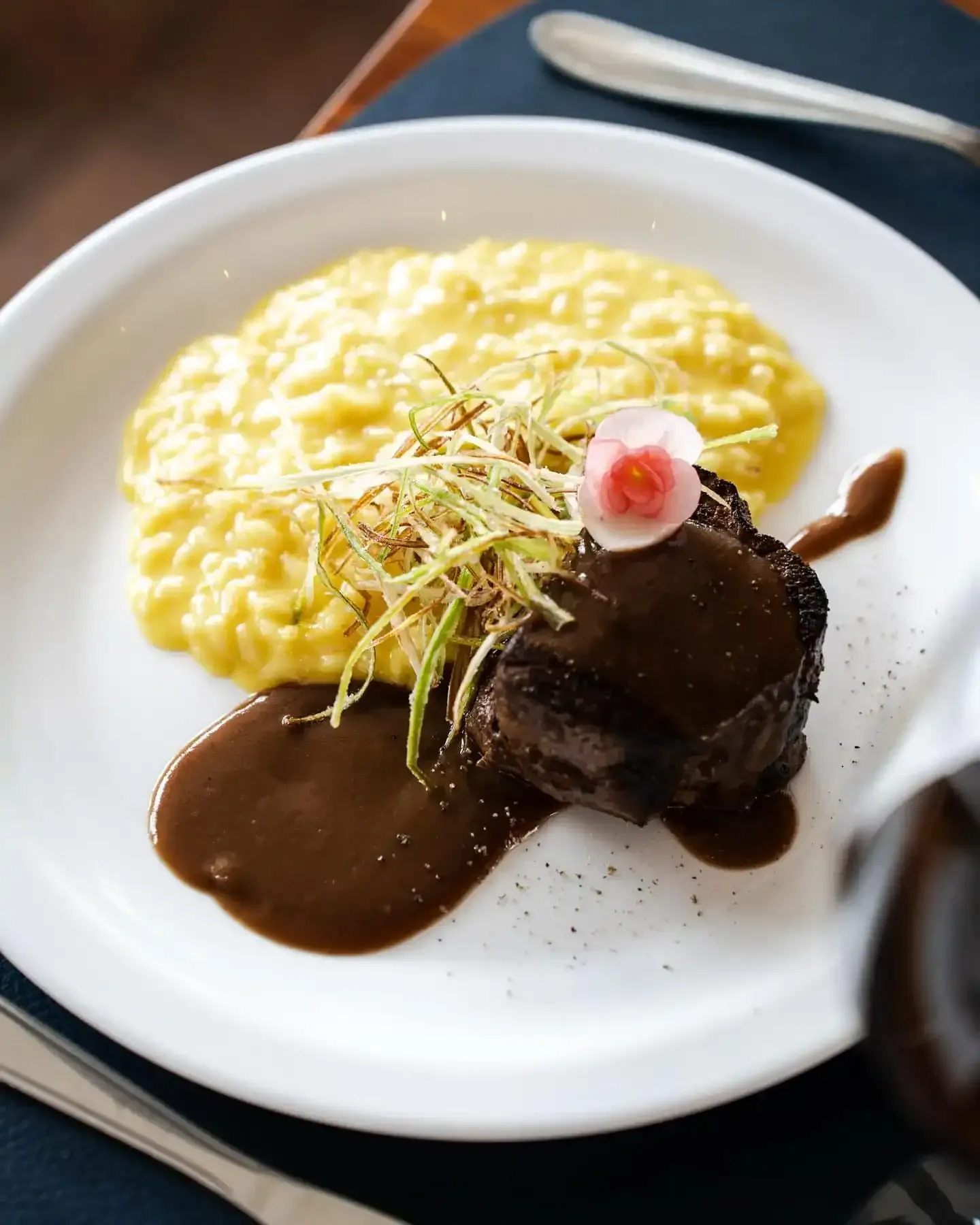 Bife com molho, risoto amarelo e guarnição em um prato branco.