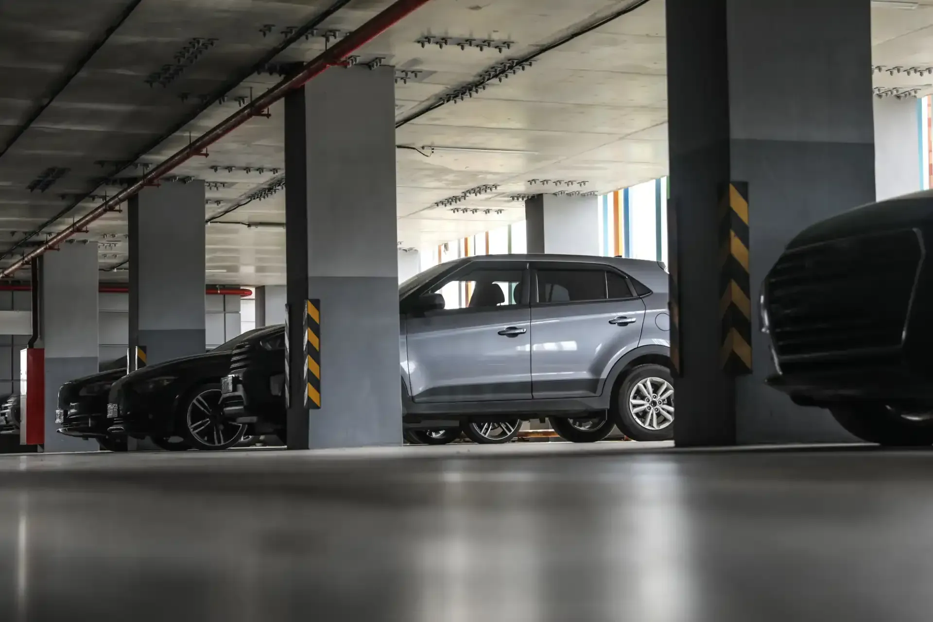 Carros estacionados em uma garagem de vários andares com colunas de concreto e tubulações suspensas.