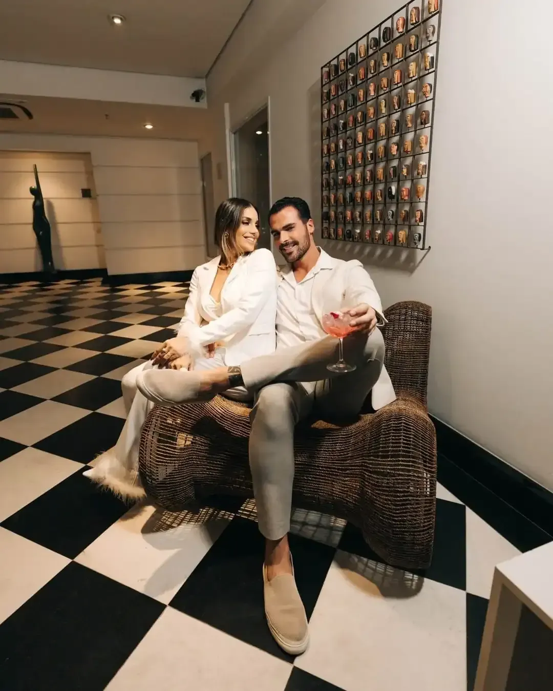 Casal sentado em cadeira ornamentada, em um corredor com piso quadriculado, olhando um para o outro.