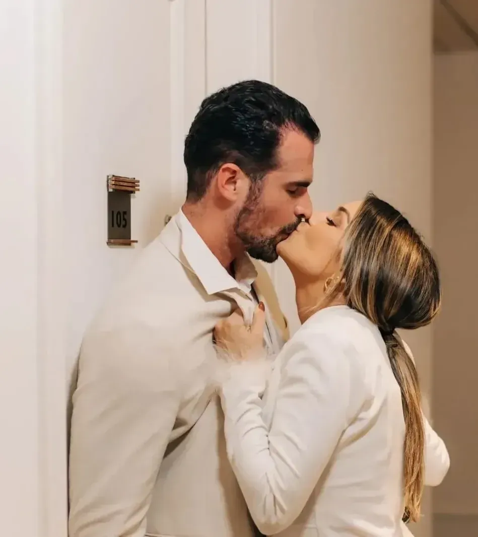 Casal se beijando no corredor, encostados um no outro. Homem de terno bege, mulher de blazer branco. Porta com o número 105.