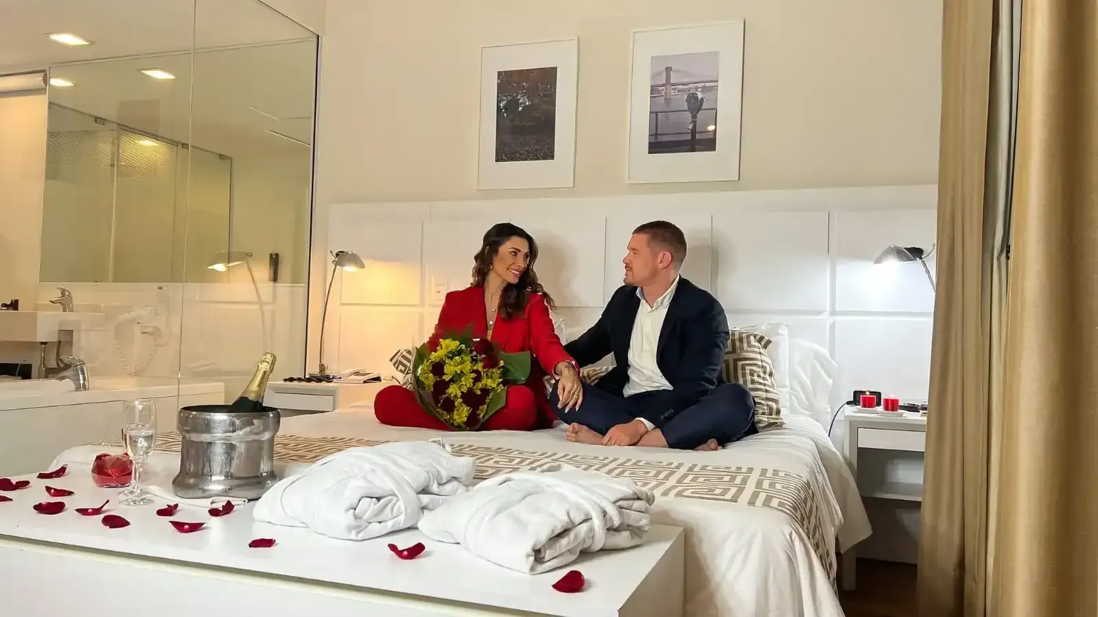 Casal sentado em uma cama de quarto de hotel, olhando um para o outro. Decoração em vermelho e branco, champanhe, flores.