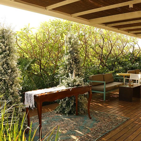 Área de estar no pátio com deck de madeira, mesa, tapete e folhagem verde ao fundo.