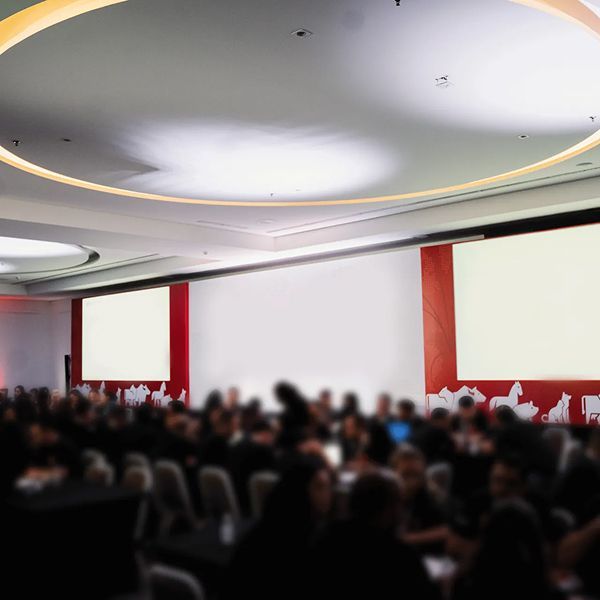 Público em sala de conferências; telas brancas, detalhes em vermelho e teto curvo.