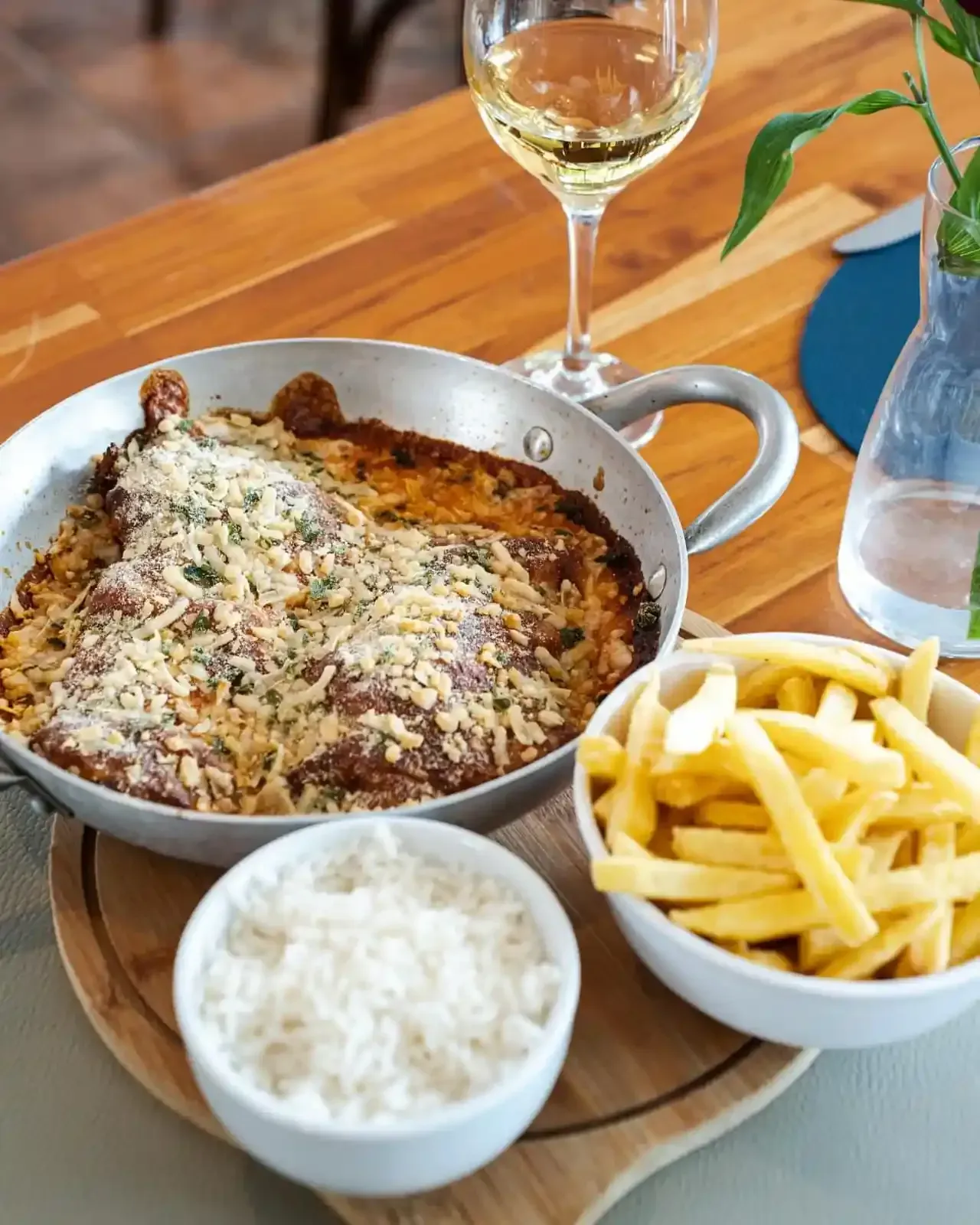 Prato de frango na frigideira com batatas fritas, arroz e vinho branco sobre uma mesa de madeira.
