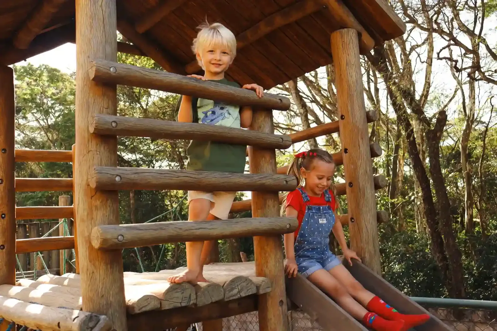 Parque infantil com escorregador, casinha de brincar e gangorra sobre tijolos vermelhos, ao lado de um edifício com mesas ao ar livre sob um toldo.