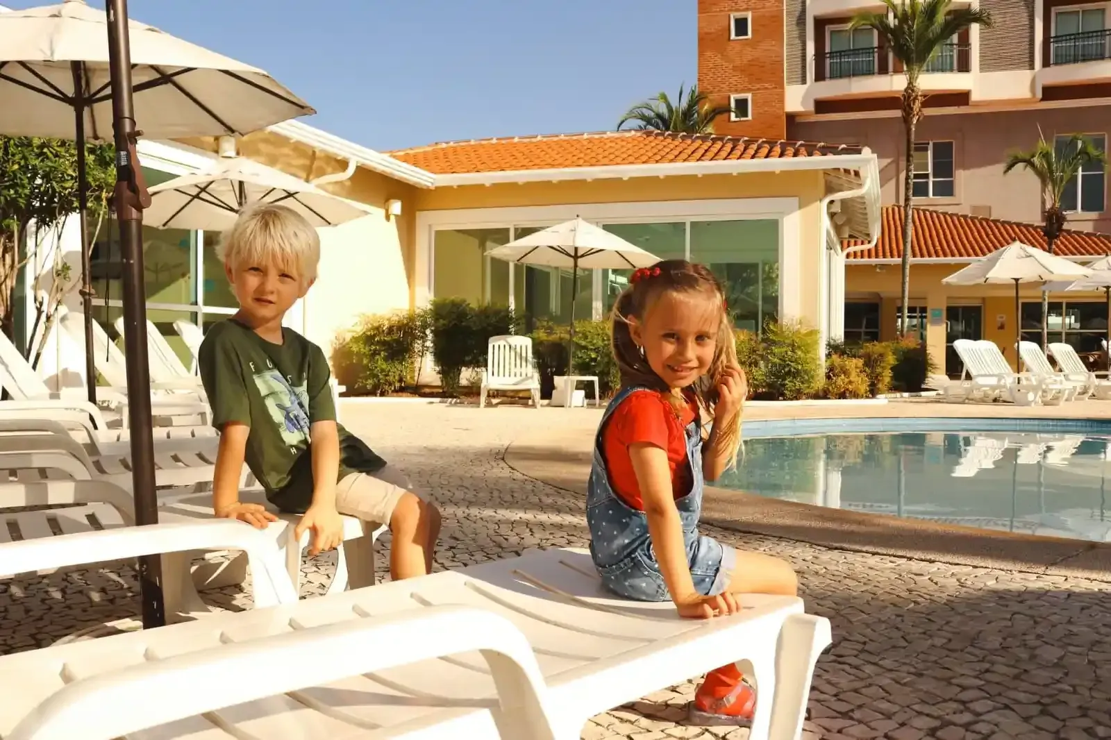 Duas crianças sorrindo à beira de uma piscina, sentadas em espreguiçadeiras em frente a um prédio com guarda-sóis.