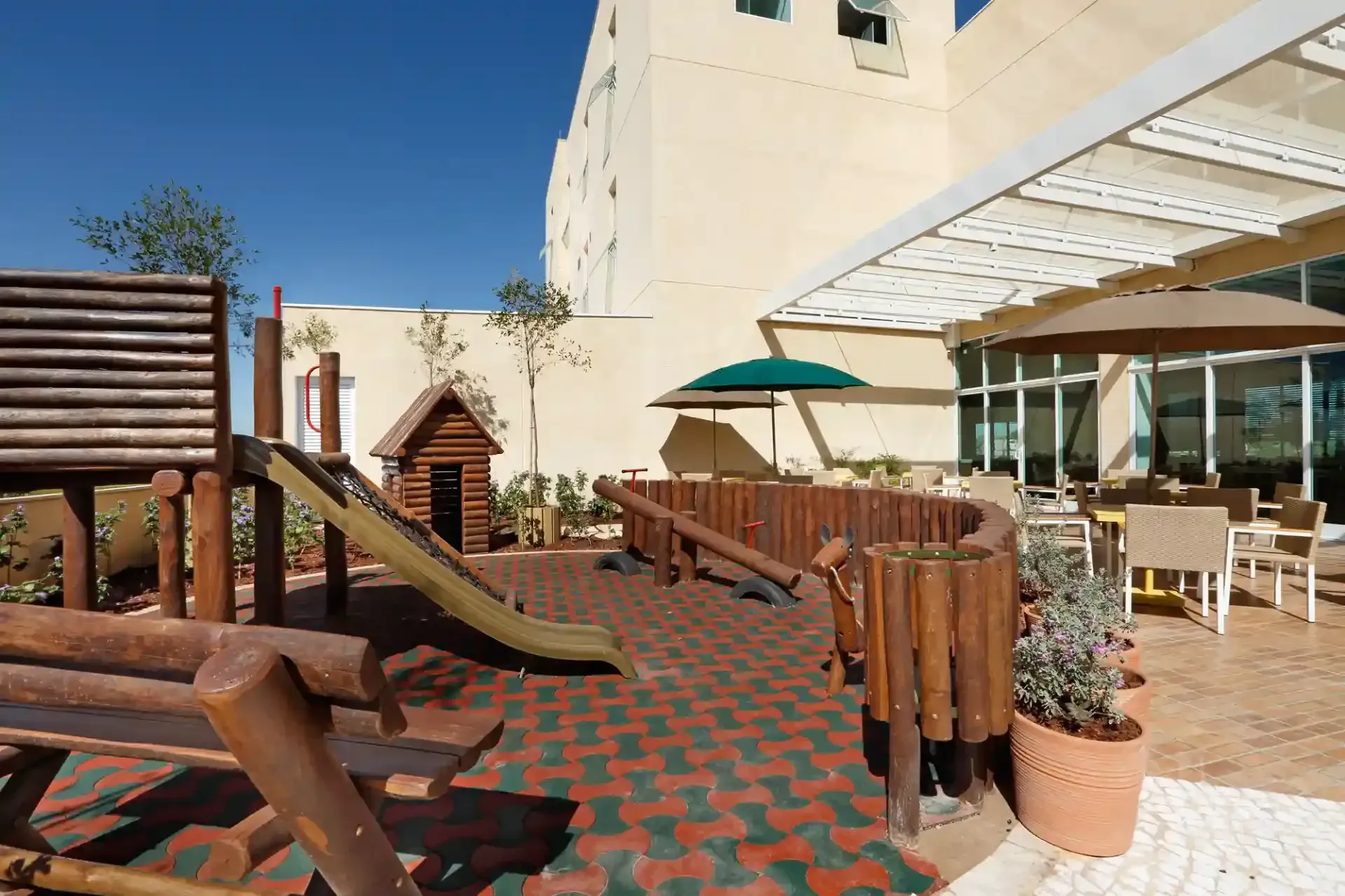 Parque infantil com estruturas de madeira, escorregador e cabana, ao lado de um edifício e área de estar sombreada.