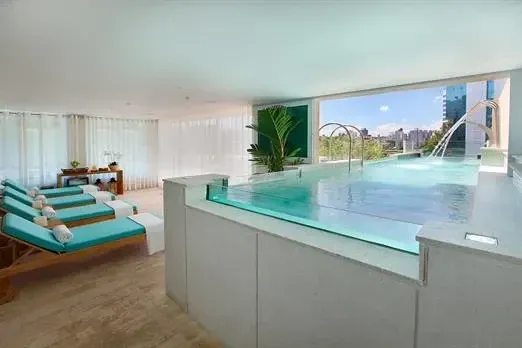 Spa interno com piscina, espreguiçadeiras e uma grande janela com vista para a cidade.