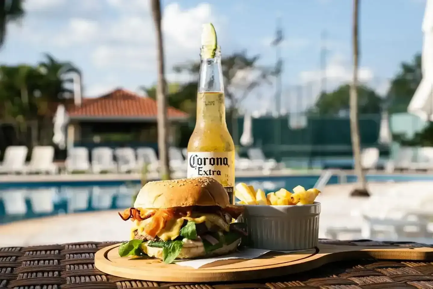 Hambúrguer, batatas fritas e cerveja Corona numa tábua de madeira junto à piscina.