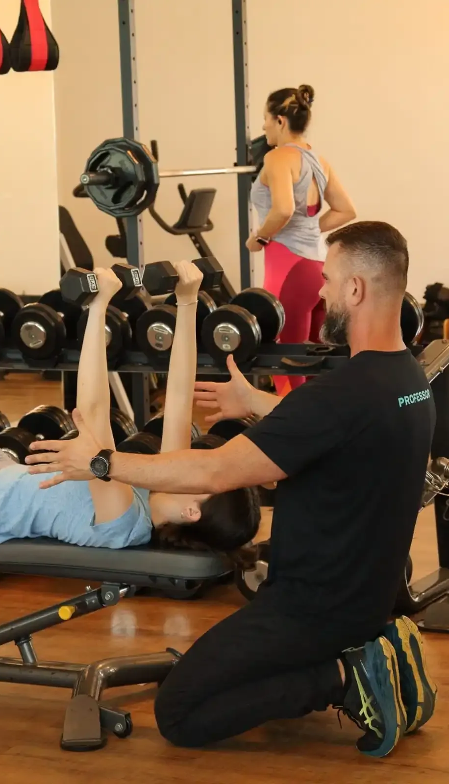 Instrutor auxiliando cliente com exercício para as pernas na academia. Outra pessoa observando.