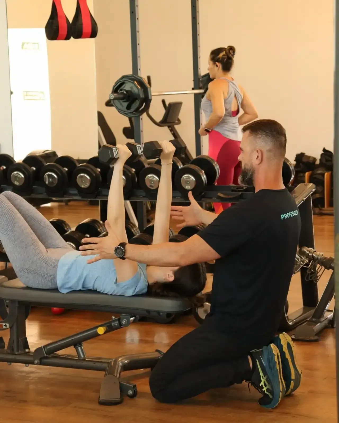 Mulher realizando supino com halteres sob a orientação do treinador. Ambiente de academia com outra pessoa ao fundo.
