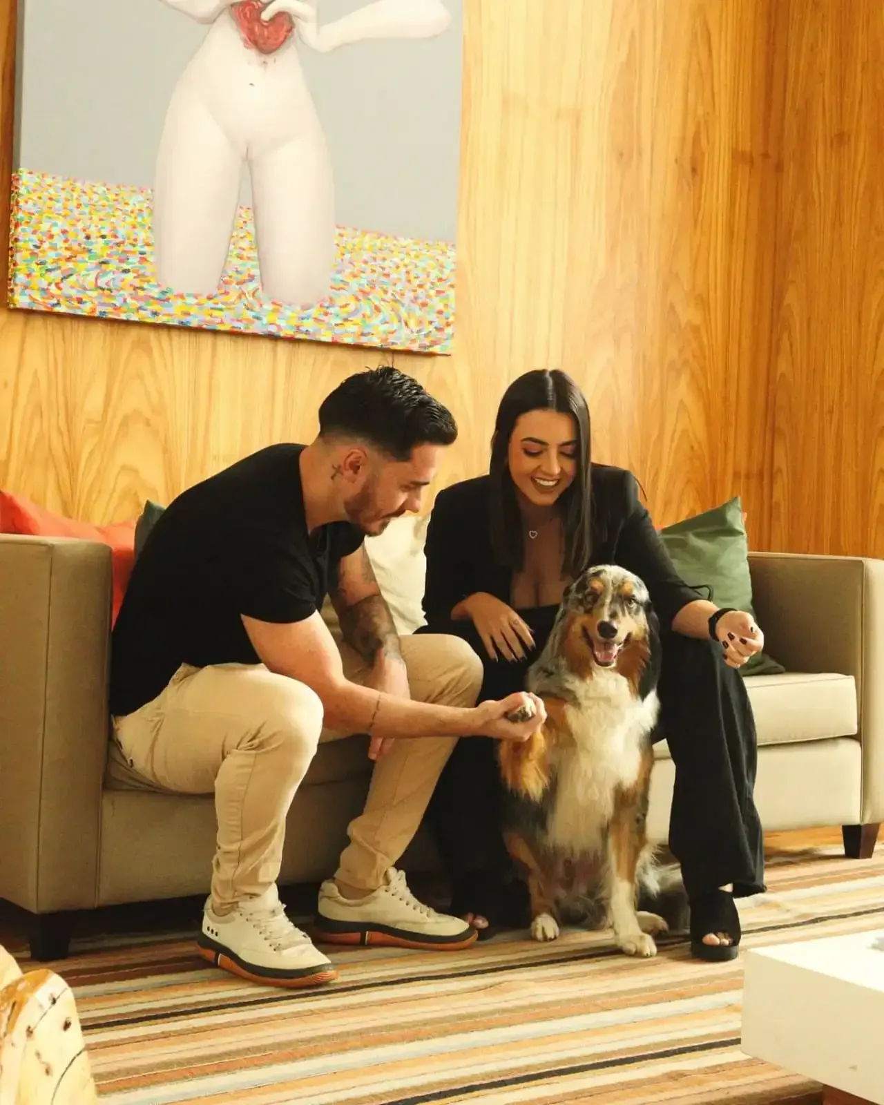 Casal no sofá com cachorro, interagindo. Interior moderno com obras de arte.