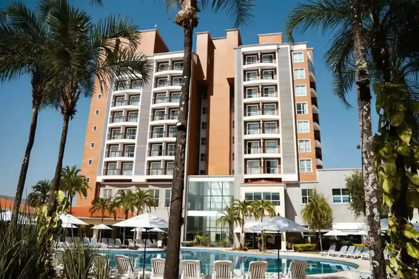 Prédio do hotel com varandas, palmeiras e piscina.