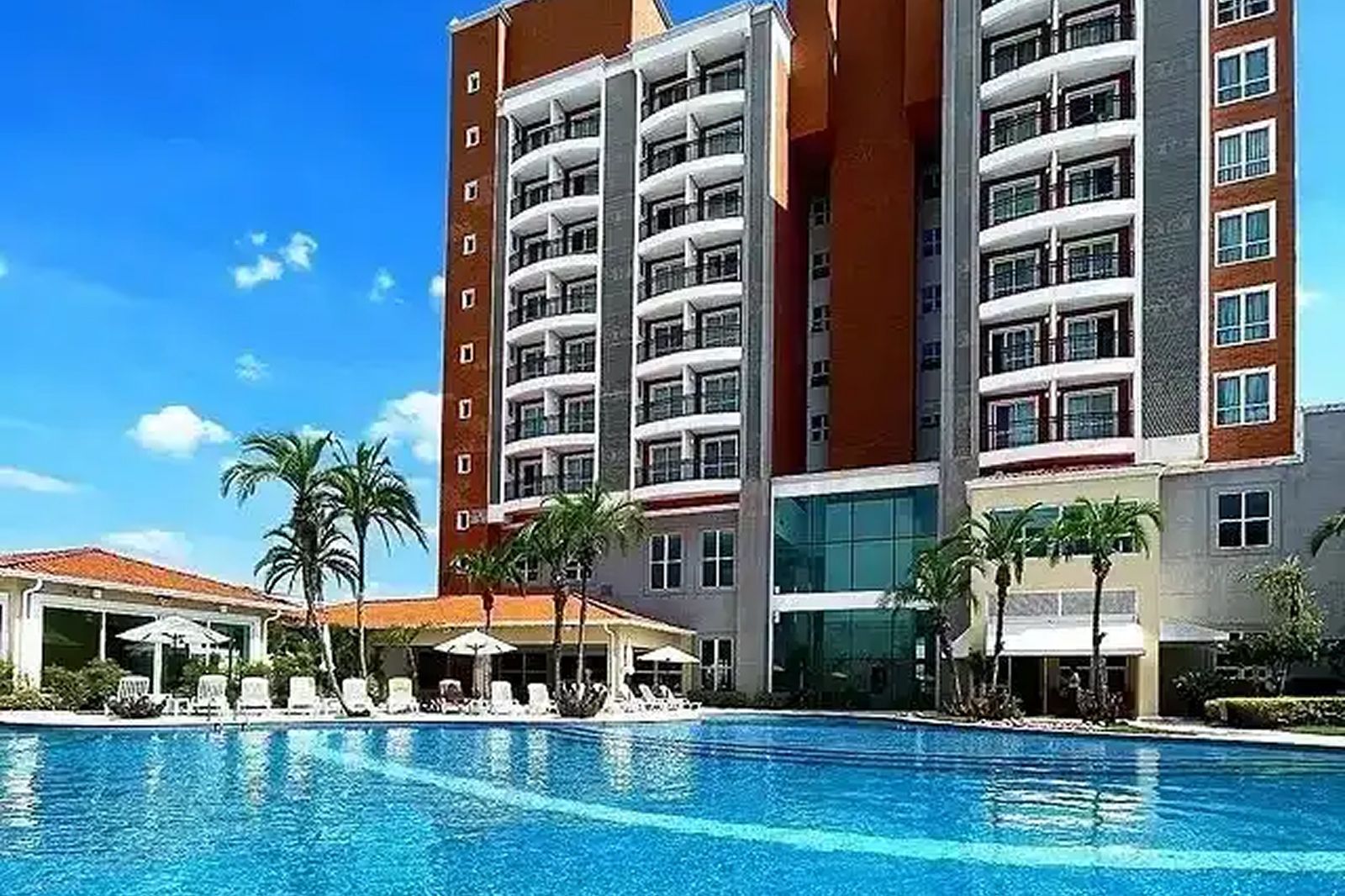 Área de descanso à beira da piscina com cadeiras vermelhas, guarda-sóis brancos e piscina revestida com azulejos azuis.