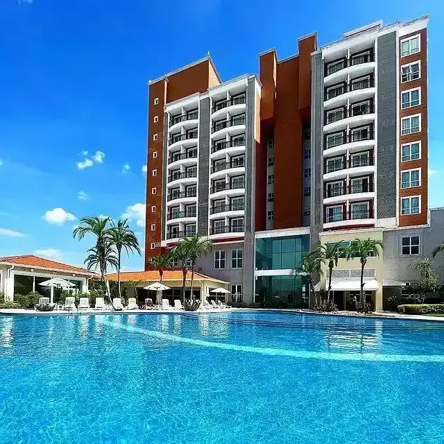 Exterior do hotel com piscina; a água azul reflete o edifício, as palmeiras e o céu ensolarado.