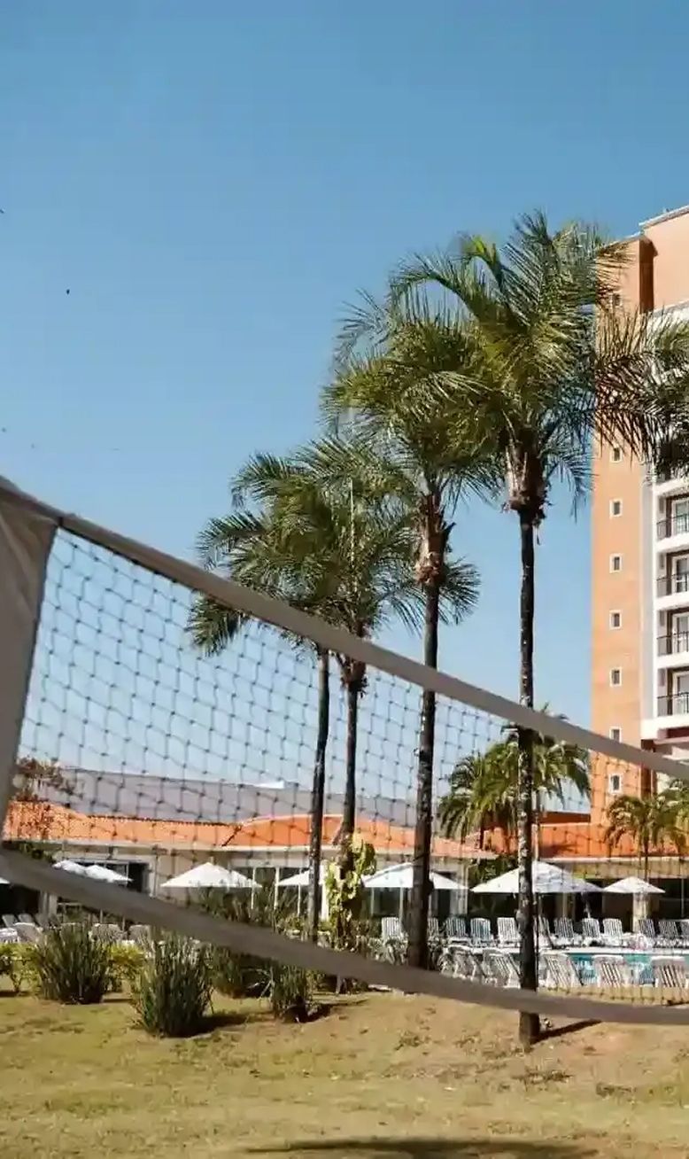 Campo de futebol com gol em primeiro plano e prédios ao fundo. Grama verde e cerca preta.