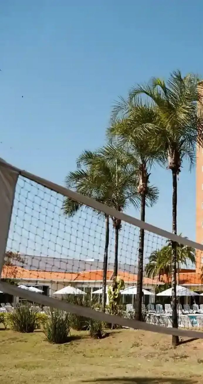 Campo de futebol com gol em primeiro plano e prédios ao fundo. Grama verde e cerca preta.