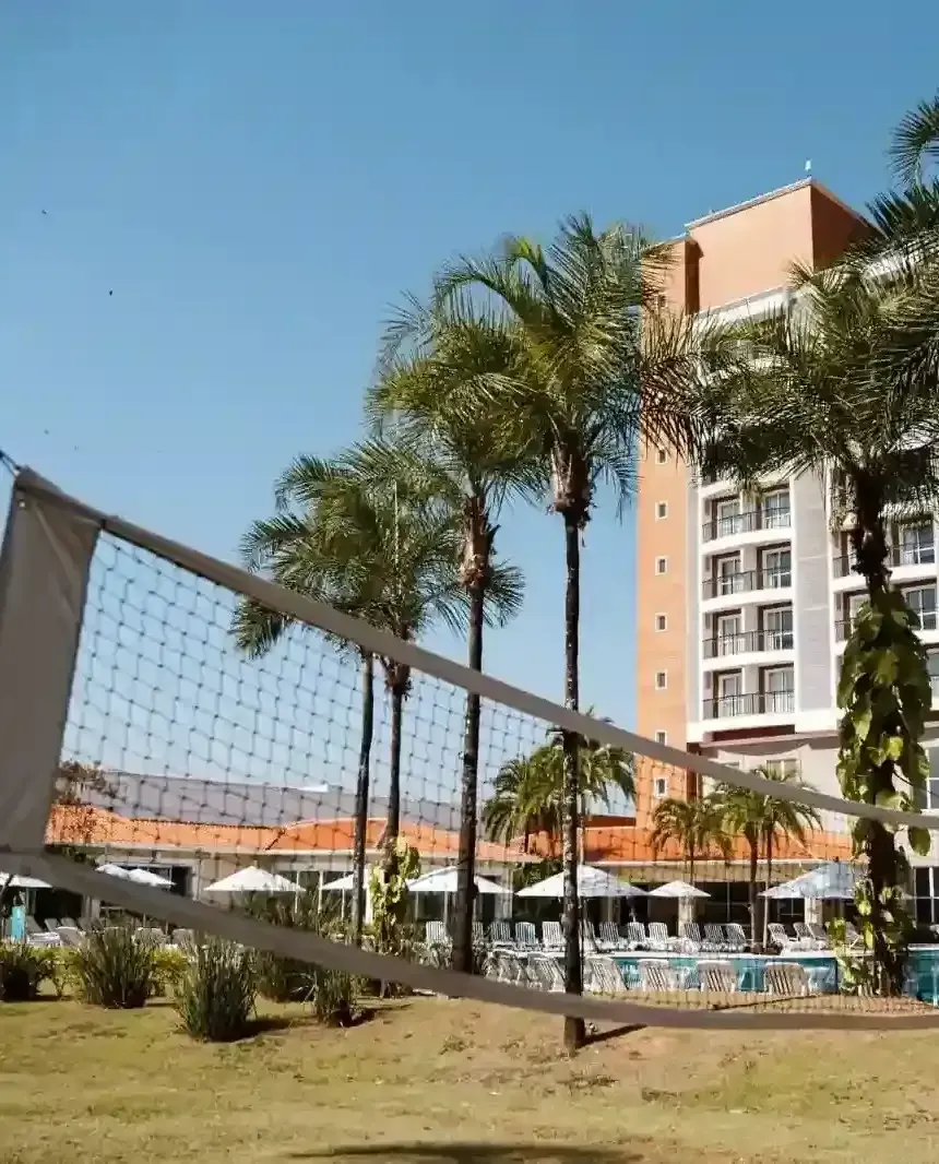Rede de vôlei em frente a um hotel com palmeiras, piscina e céu azul.