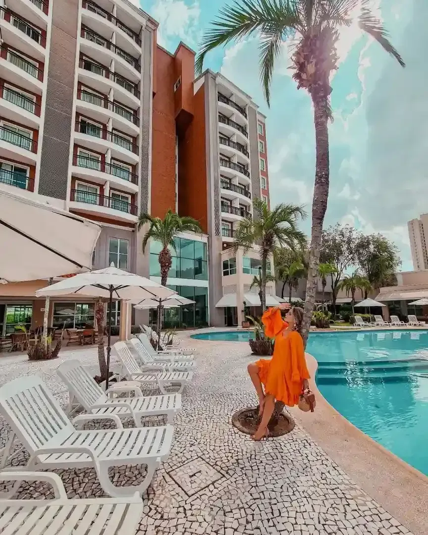Mulher de vestido laranja sentada à beira da piscina em frente a um hotel com palmeiras, em um dia ensolarado.