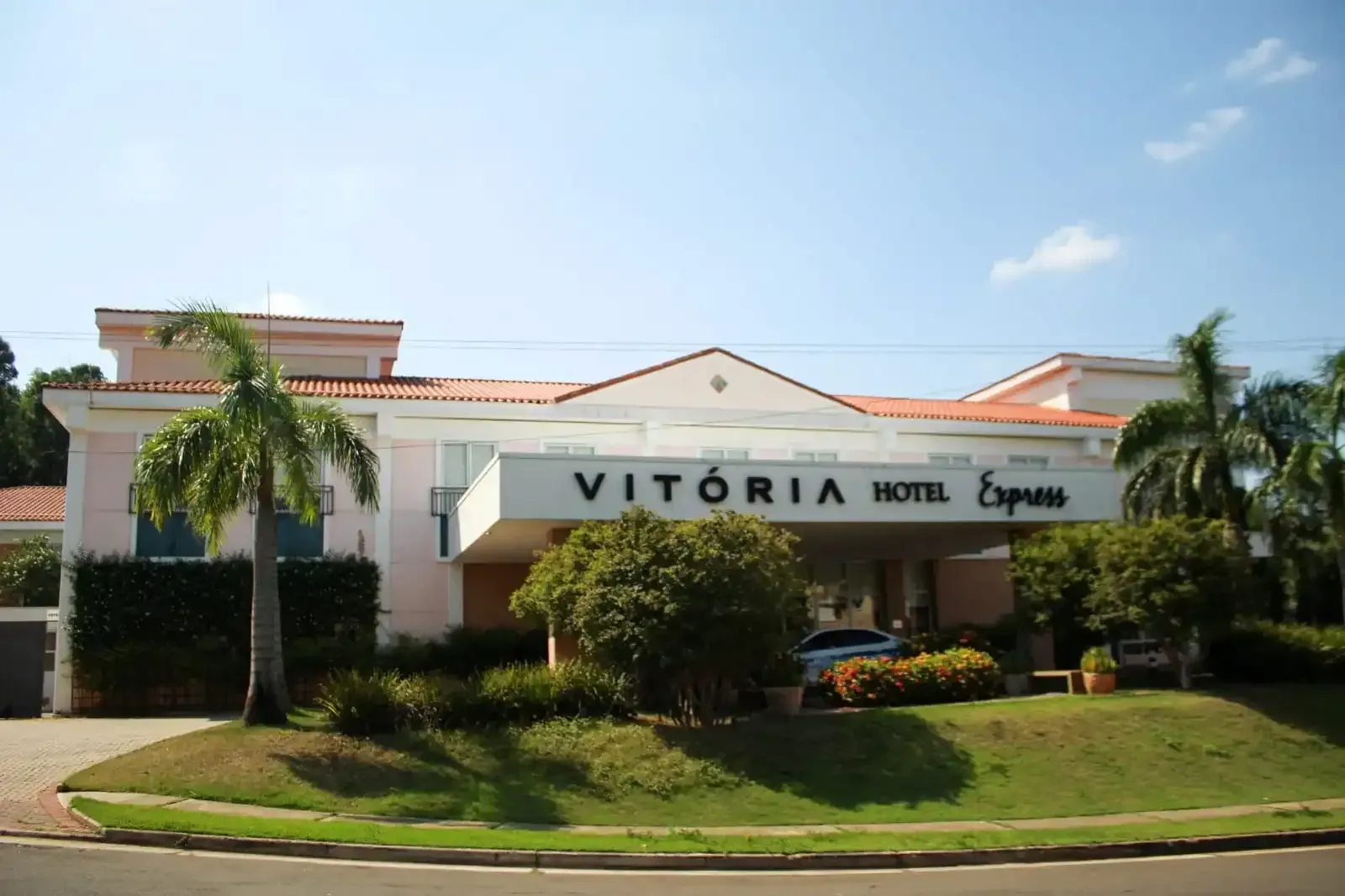 Vista exterior do Vitoria Hotel Express. Prédio branco, placa grande, palmeiras, gramado verde, céu limpo.