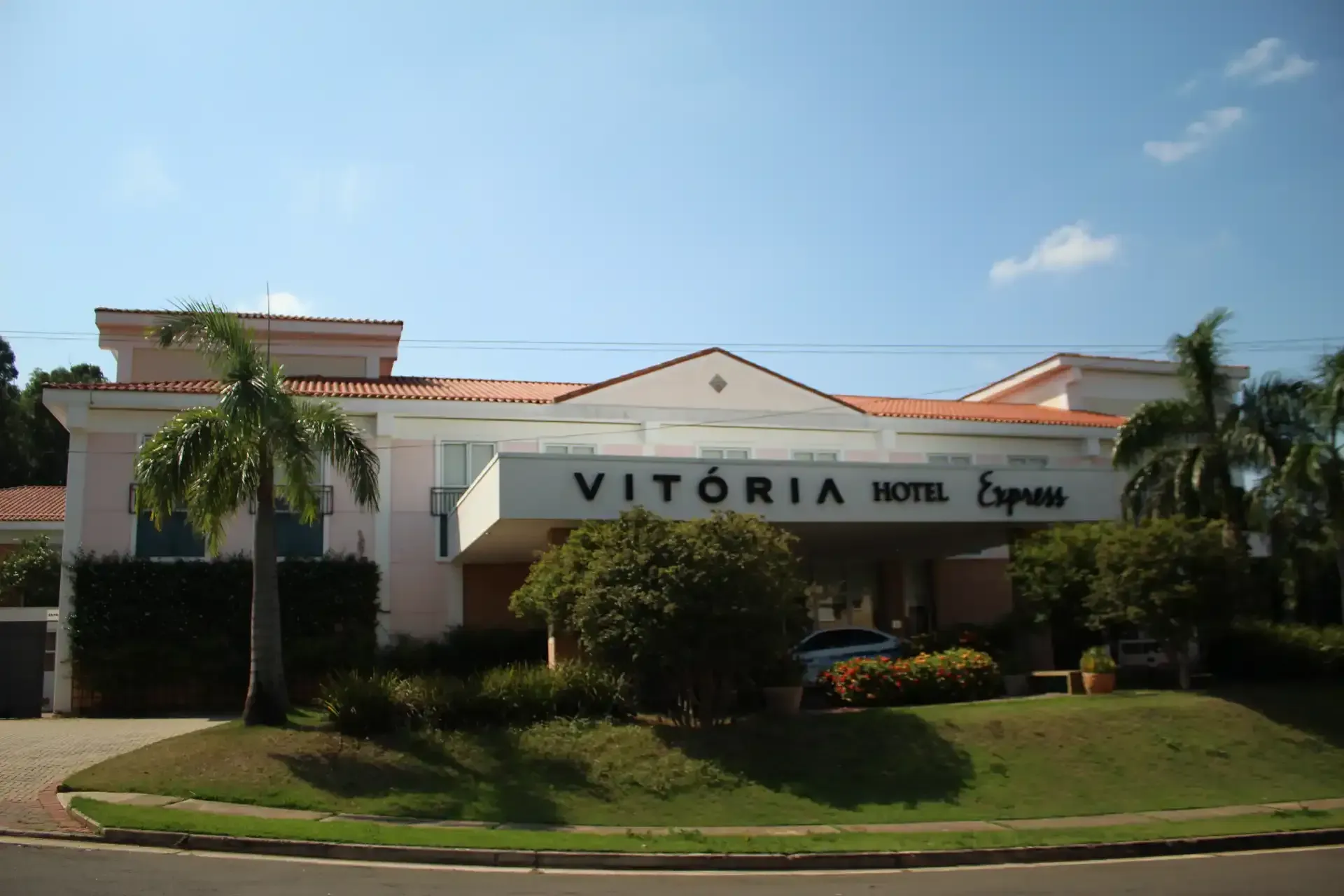 O Vitoria Hotel Express, um edifício de cor clara com uma placa, palmeiras e gramado, sob um céu azul.