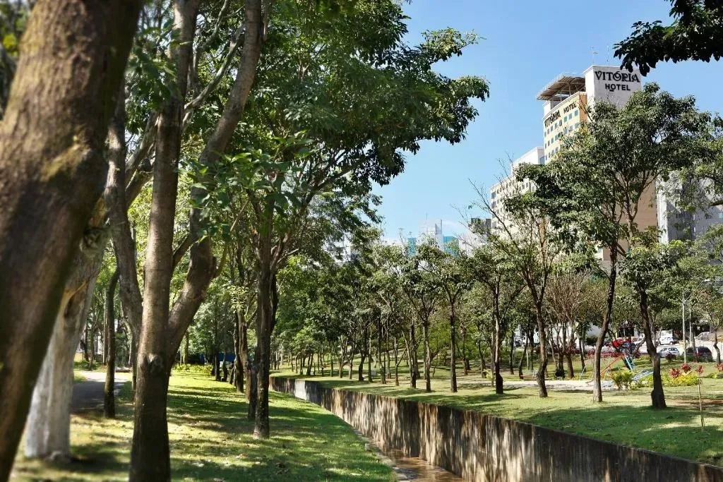 Árvores margeiam um curso d'água em um parque, com um prédio alto visível ao fundo sob um céu azul.