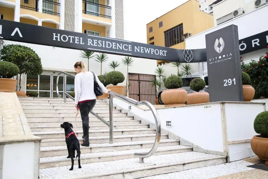 Mulher com cachorro sobe as escadas da entrada do hotel. Placa "Hotel Residence Newort" visível.