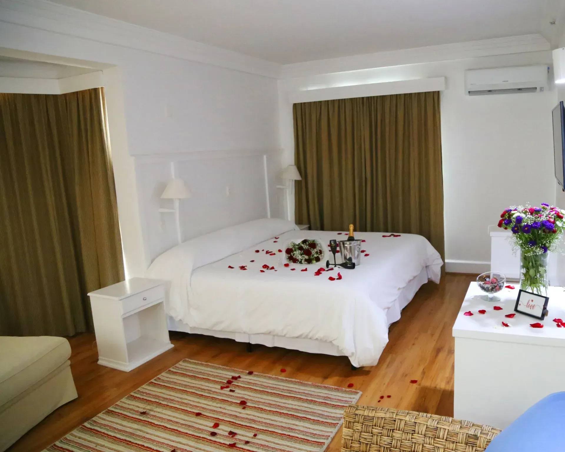 um quarto de hotel com uma cama cheia de pétalas de rosas, um buquê de rosas, 2 taças e uma garrafa de espumante