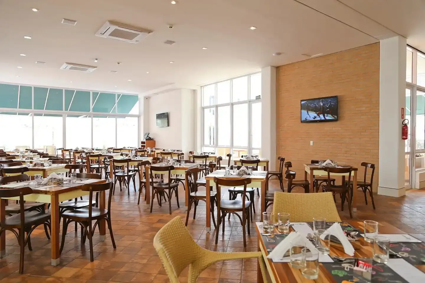 Interior de restaurante com mesas, cadeiras e grandes janelas. Tons de marrom e branco.