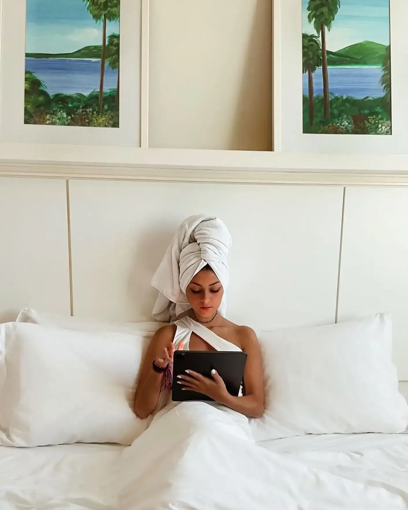 Mulher de toalha e roupão brancos deitada na cama, usando um tablet. Quadros estão pendurados acima da cabeceira.