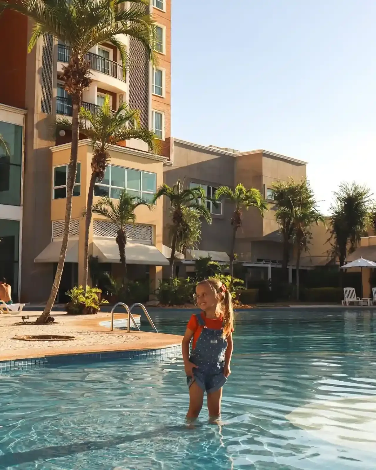 Uma garota de macacão jeans está em pé na piscina, sorrindo, em um resort tropical.