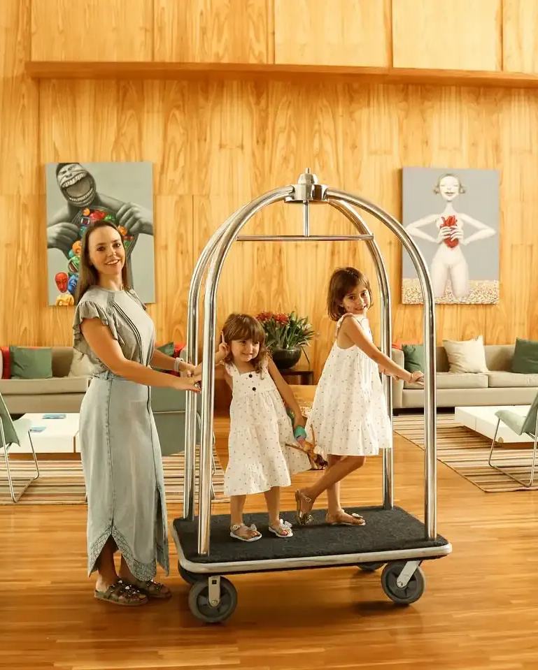 Uma mulher empurra um carrinho de bagagem com duas crianças dentro. Hall de entrada de hotel com obras de arte nas paredes.