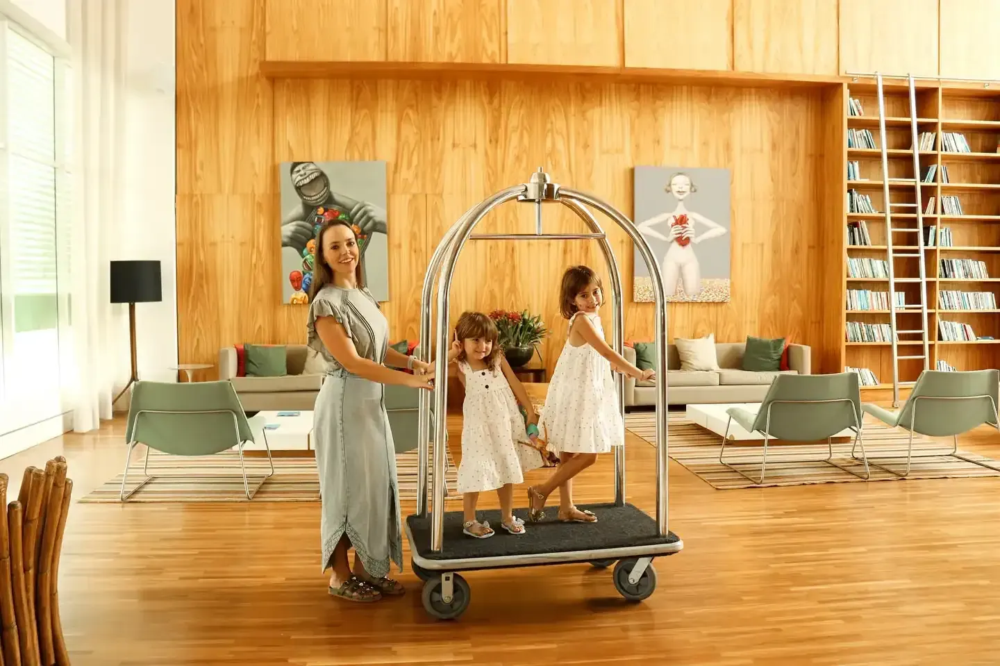 Uma mulher empurra um carrinho de bagagem com duas crianças em um saguão moderno e iluminado, decorado com obras de arte e painéis de madeira.