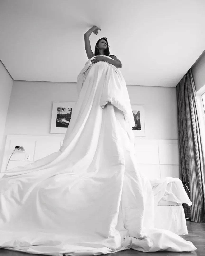 Mulher em pé sobre uma cama, coberta por um grande lençol branco, com o braço levantado. Em um quarto.