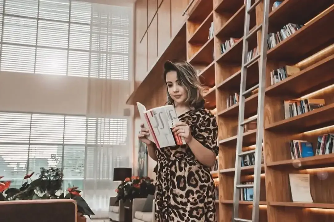 Mulher lendo um livro em uma biblioteca. Ela está usando um vestido com estampa de leopardo, com estantes de livros e uma escada ao fundo.
