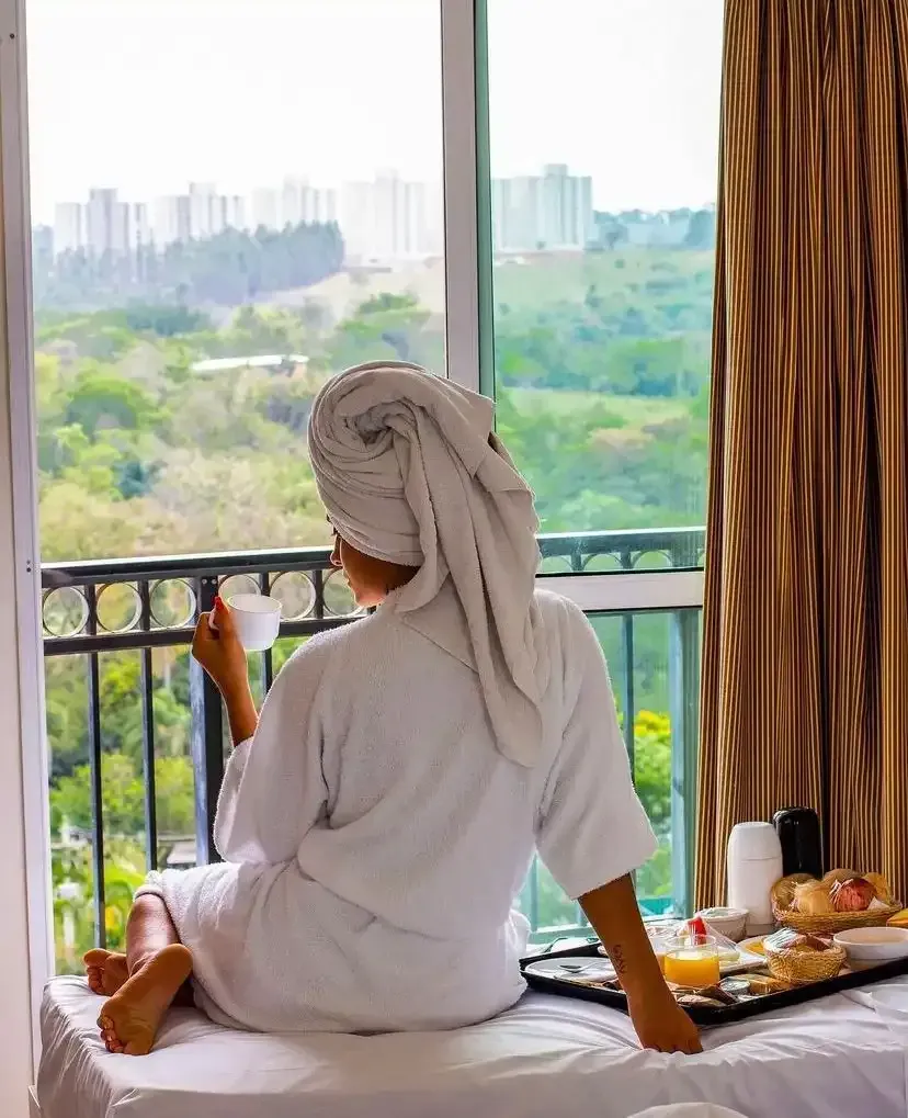 Pessoa de roupão e toalha olhando pela janela, segurando uma caneca. Bandeja de café da manhã na cama. Vista da cidade.