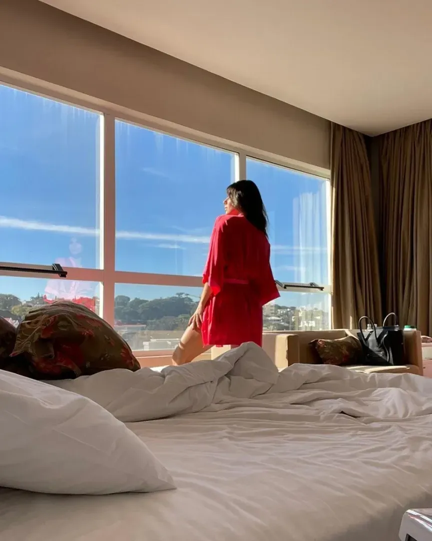 Mulher de robe vermelho olhando pela grande janela com céu azul. Cama em primeiro plano.