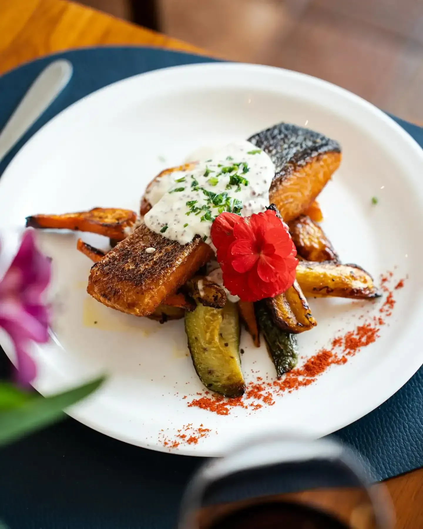 Prato de salmão com legumes assados, molho cremoso e flor vermelha como decoração, servido em prato branco.