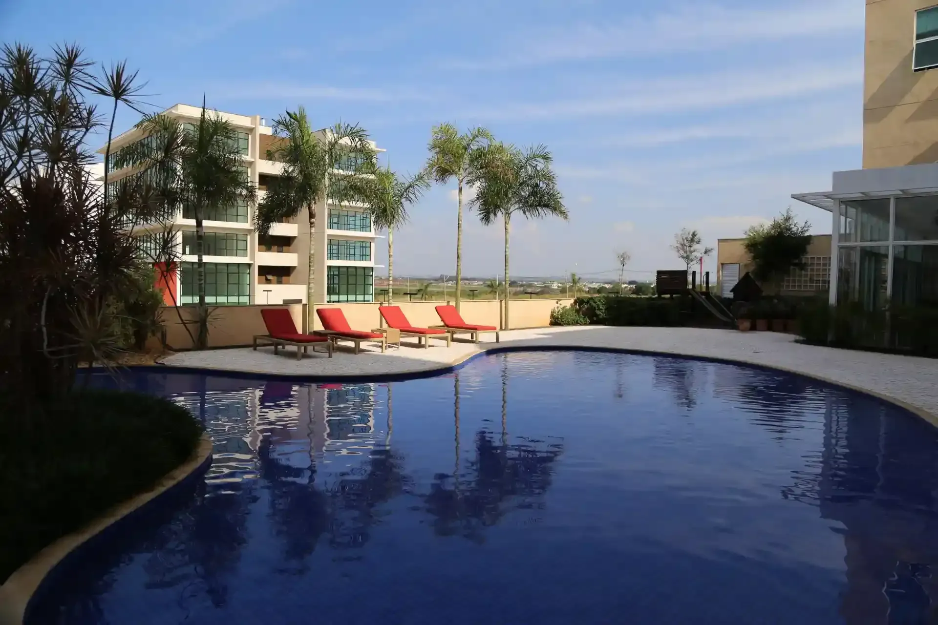 Piscina com espreguiçadeiras, árvores e edifícios sob um céu azul.