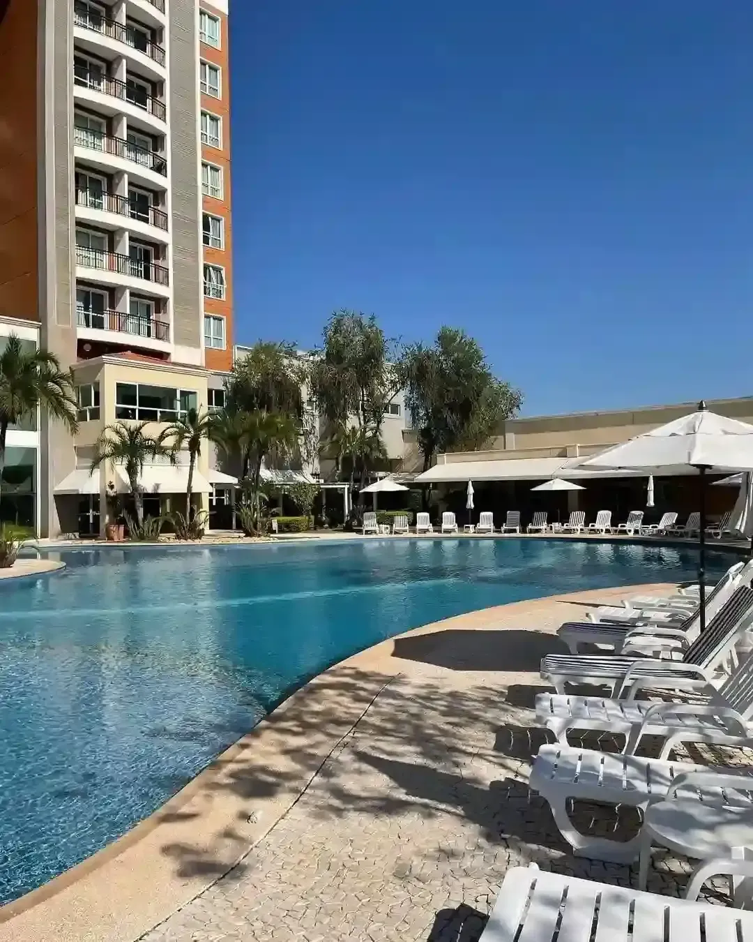 Piscina em um resort com espreguiçadeiras, guarda-sóis e um prédio alto em um dia ensolarado.