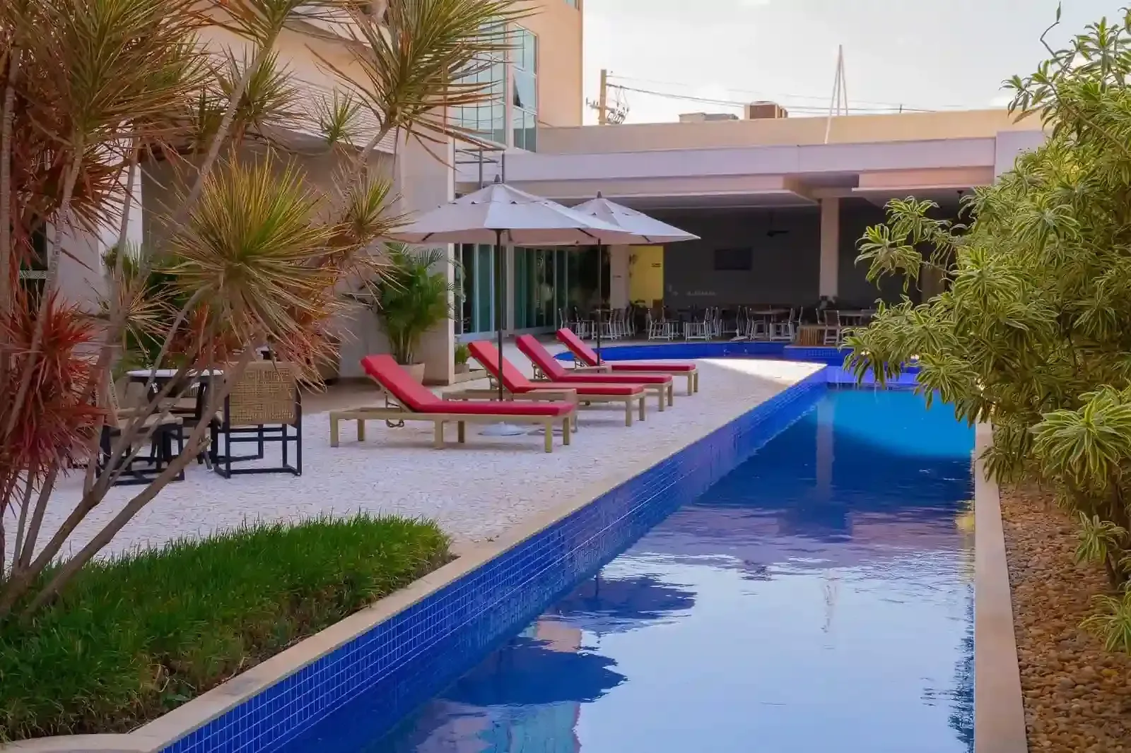 Área de descanso à beira da piscina com cadeiras vermelhas, guarda-sóis brancos e piscina revestida com azulejos azuis.