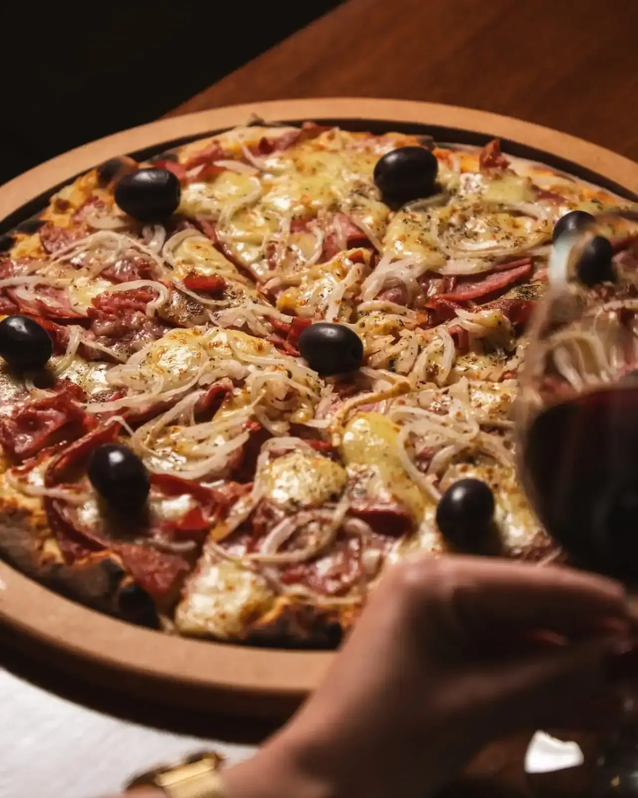 Pizza servida em uma tábua de madeira com azeitonas, cebolas e carne. Uma mão segurando uma taça de vinho tinto.